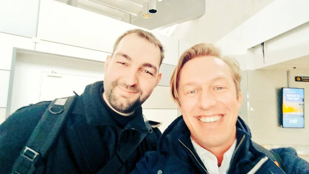 Two men smile together for a selfie indoors.