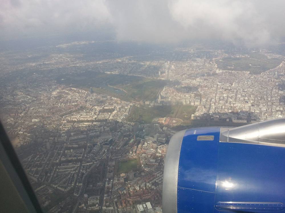 Aerial view of London's dense city blocks from the plane, wingtip and engine visible below scattered clouds.