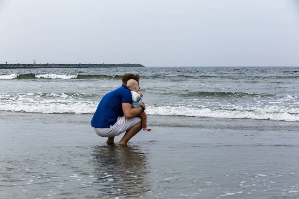 A man crouches to hug a toddler at the water's edge on a beach under a dark sky.
