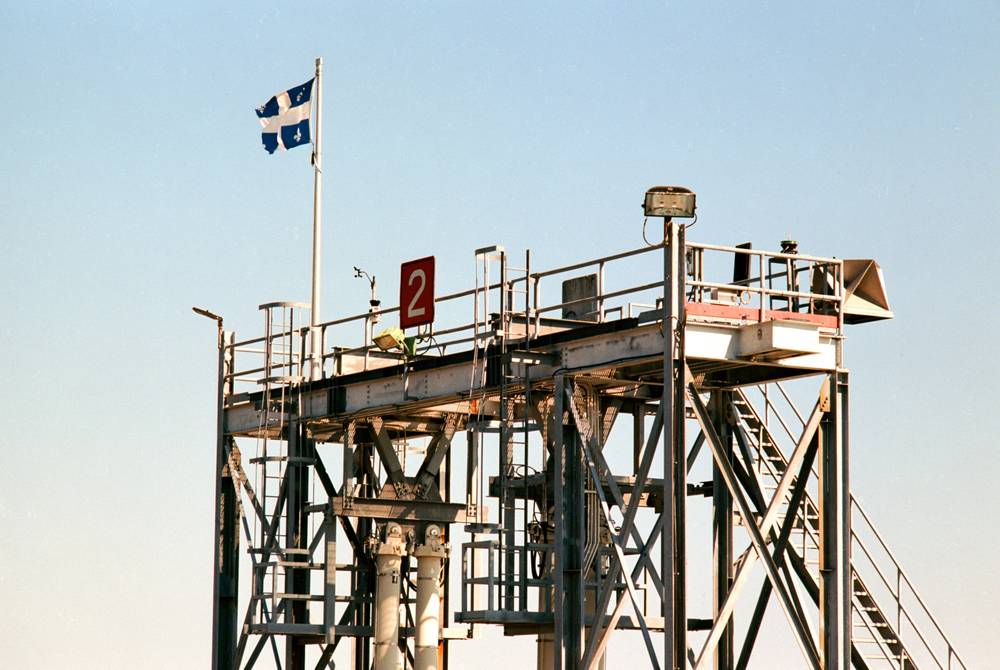 A large industrial metal structure under a clear blue sky features a Quebec flag flying from a tall pole on its upper platform.