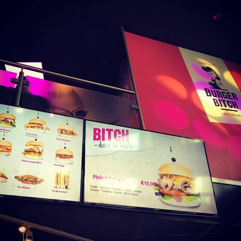 Illuminated signage for a "Burger Bitch" restaurant advertising burgers on digital menu boards.
