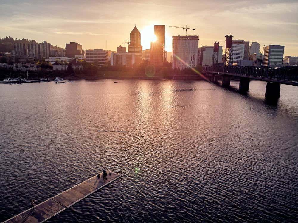 The sun sets behind Portland's downtown skyline, casting brilliant rays between the towering buildings and painting the Willamette River in molten copper tones. The iconic Hawthorne Bridge stretches across the frame while construction cranes hint at the city's ongoing growth. From Holman Dock on the Vera Katz Eastbank Esplanade, this elevated perspective captures the serene convergence of urban architecture and natural waterway during the magical golden hour.