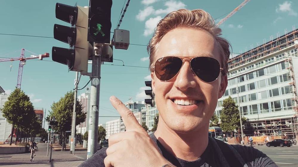 Selfie at a city street crossing, pointing at traffic lights.