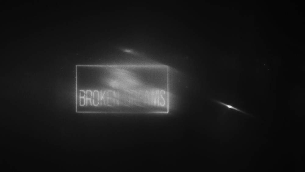 Black and white, a sign reading BROKEN DREAMS floats in the grey. The sign itself is blurred as if in motion