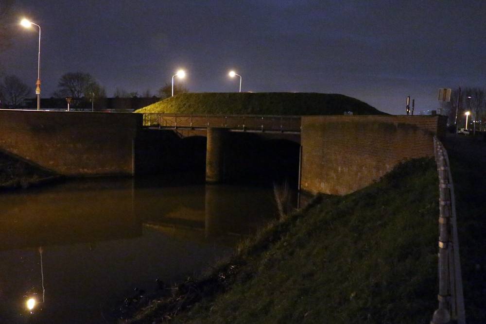 A stone bridge spans a dark canal beneath streetlights at night.