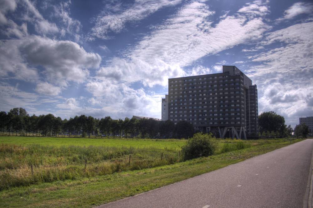 A tall dark apartment tower rising above a grassy field along a bike path under a partly cloudy sky.