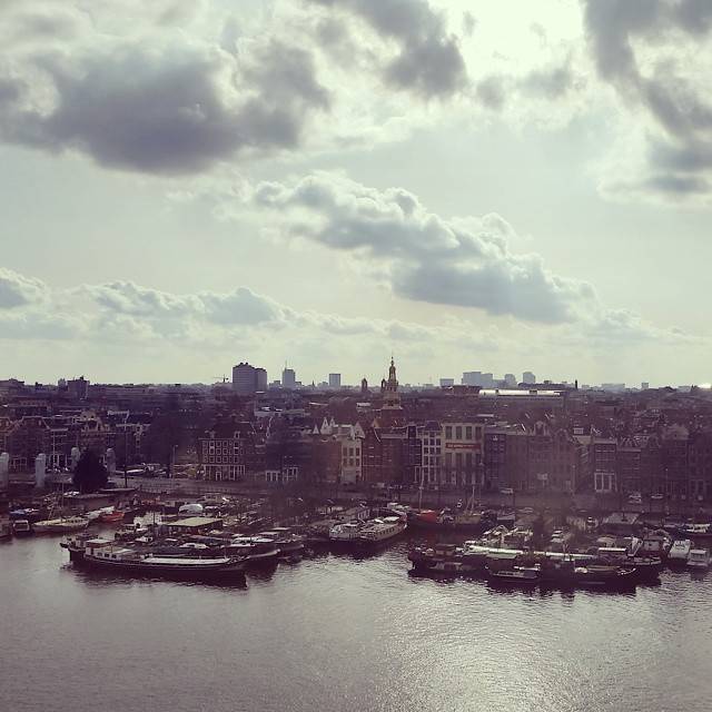 Aerial view of Amsterdam's historic harbor filled with boats, with the city skyline beyond.