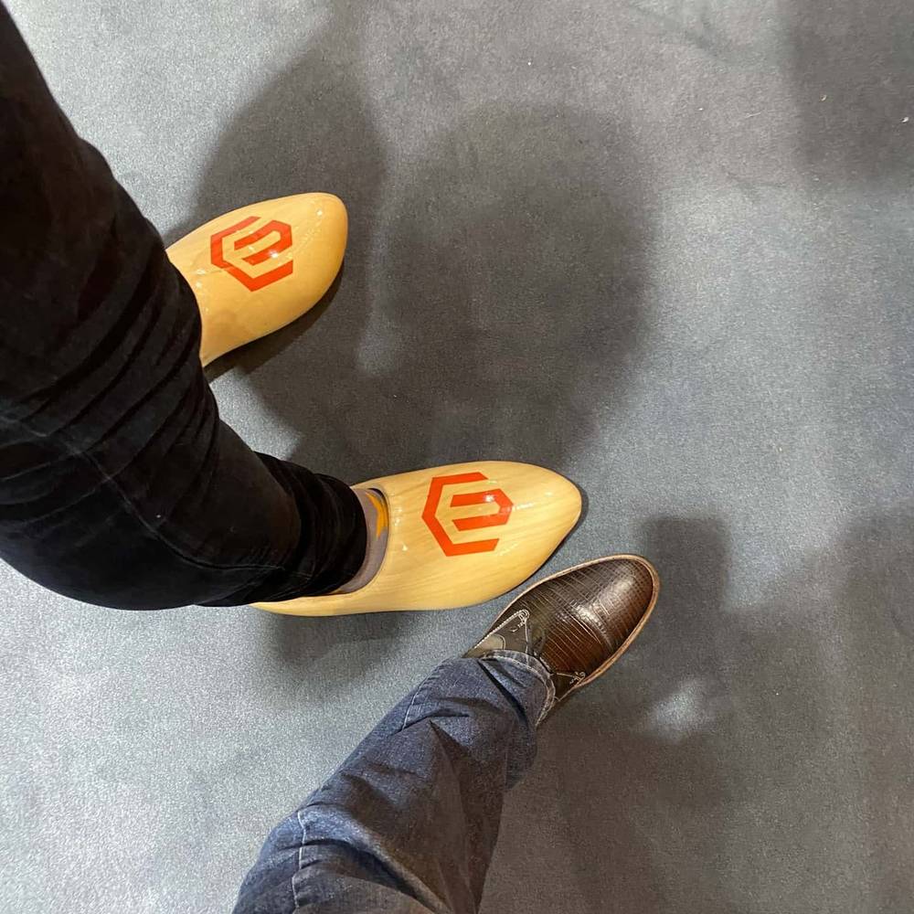Close-up of feet wearing wooden clog-style shoes with a logo, next to leather dress shoes.