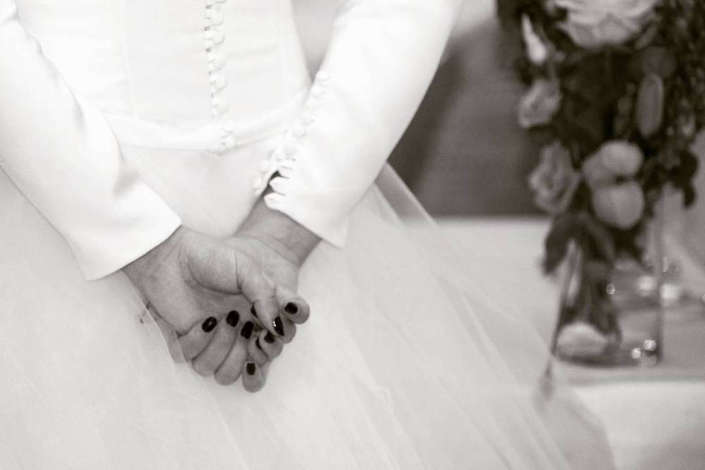 Black and white close-up of a bride and groom's clasped hands beside a flower vase.