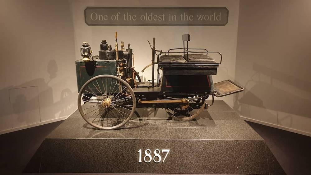 An antique horse-drawn style automobile on a museum pedestal, labeled 1887 as one of the oldest in the world.
