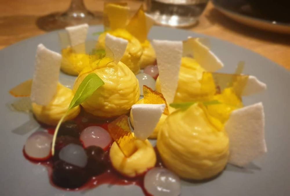 A plated dessert with yellow mousse quenelles, meringue pieces and berries.