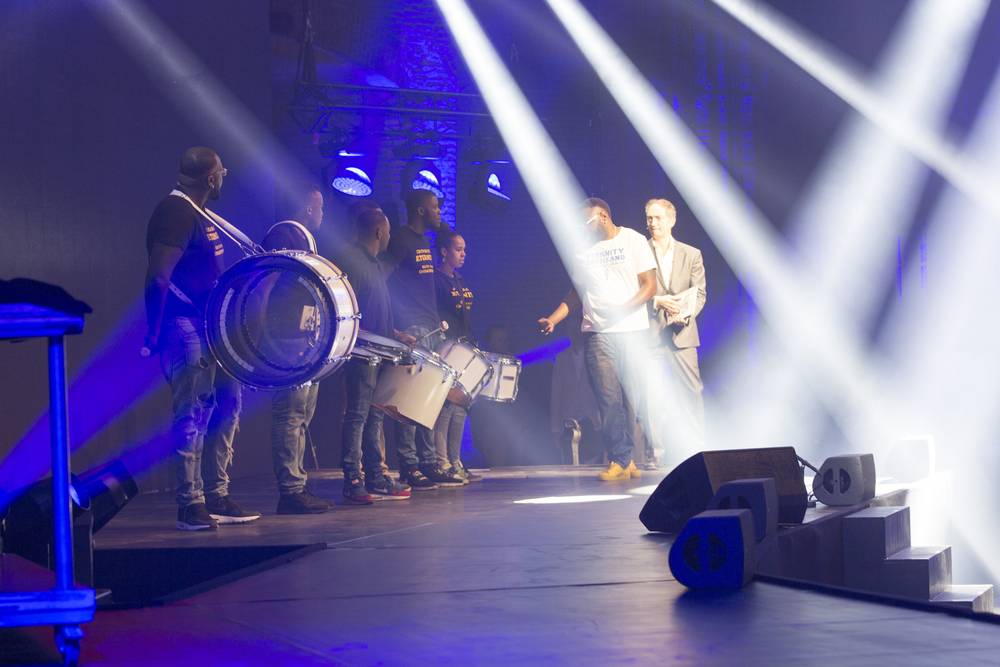 Performers with a large drum walk on stage as two men in suits look on during an opening ceremony.