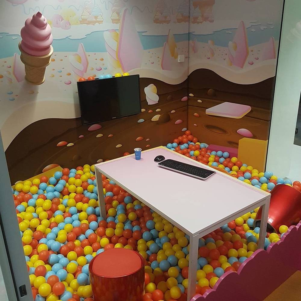An office break room with a colorful mural, a ball pit, and a desk with a keyboard.