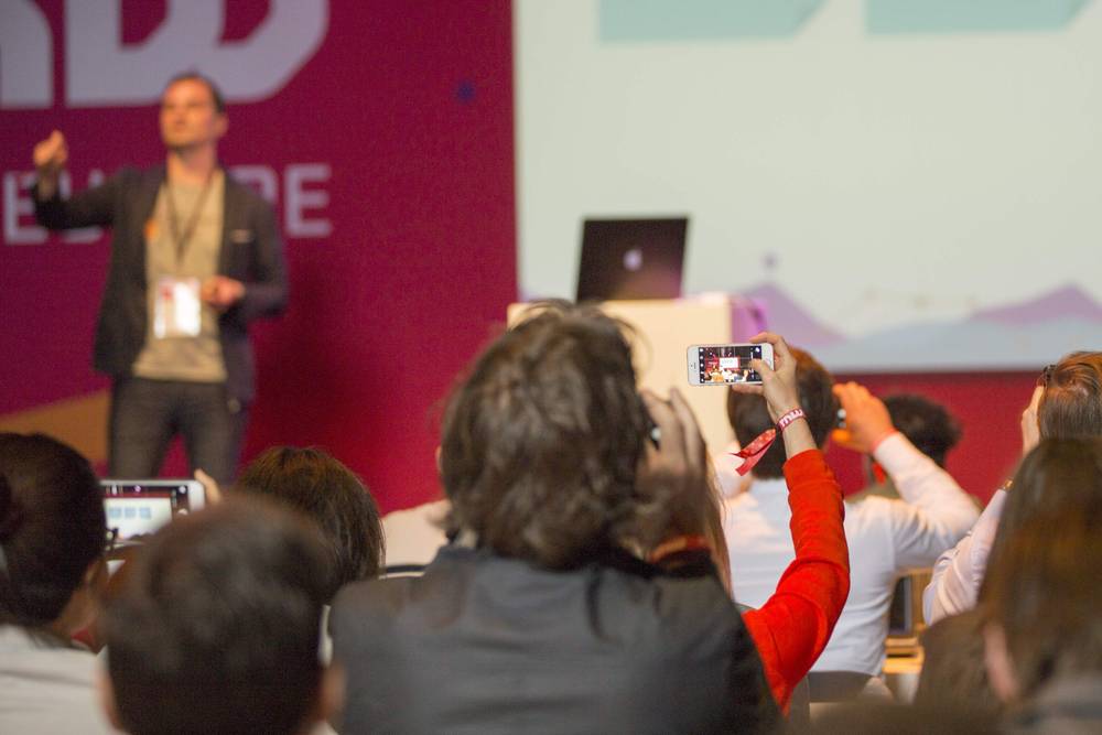 A speaker gestures on stage as an audience member raises a phone to film below.