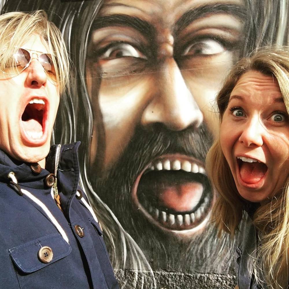 A selfie of two people making exaggerated surprised expressions in front of a large painted mural face.