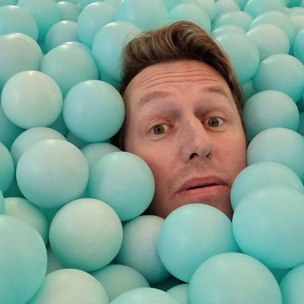 A man's face poking out from a pile of pale turquoise ball-pit balls, with a surprised expression.