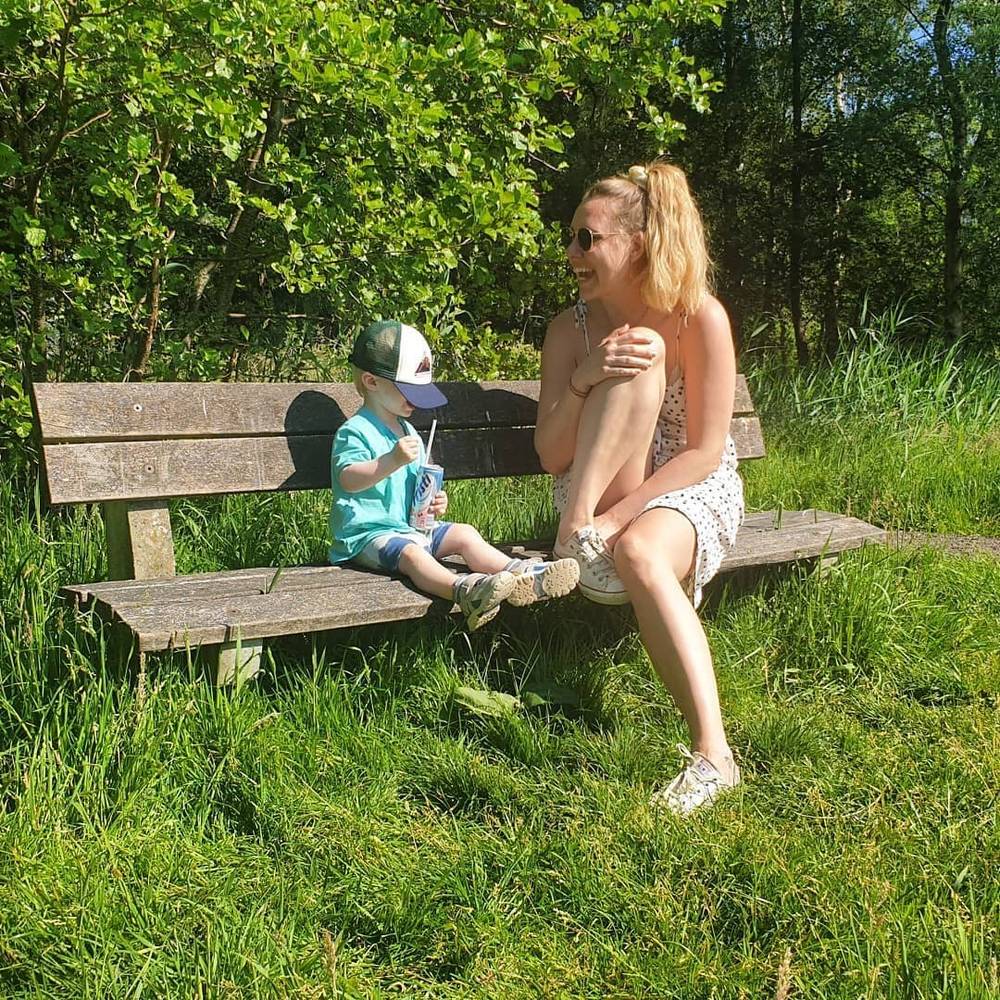 A woman and a toddler sitting together on a wooden bench in a sunny grassy area surrounded by trees.