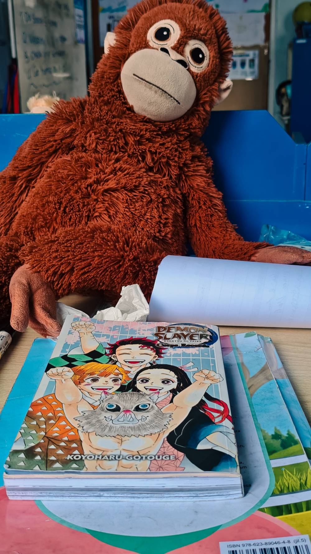 In the front, a colorful Demon Slayer manga cover sits on a stack of books, featuring the anime squad looking hyped and ready to fight demons.
Right behind it, a giant, fluffy orange orangutan plushie is slouched against a blue backdrop. He has huge, blank eyes staring into space with absolutely zero thoughts.
It perfectly captures the vibe of an anime team ready to save the world, backed up by a giant monkey having an existential crisis.