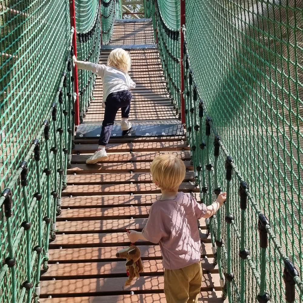 Two children walking hand over hand across a suspended net bridge at a playground.