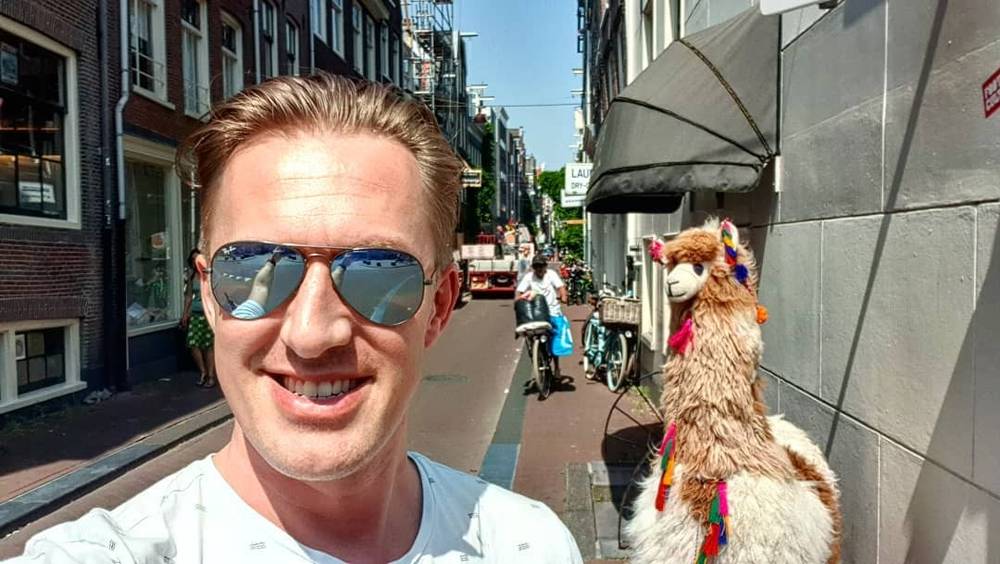 A man takes a selfie on a street beside a decorated llama figure outside a shop.