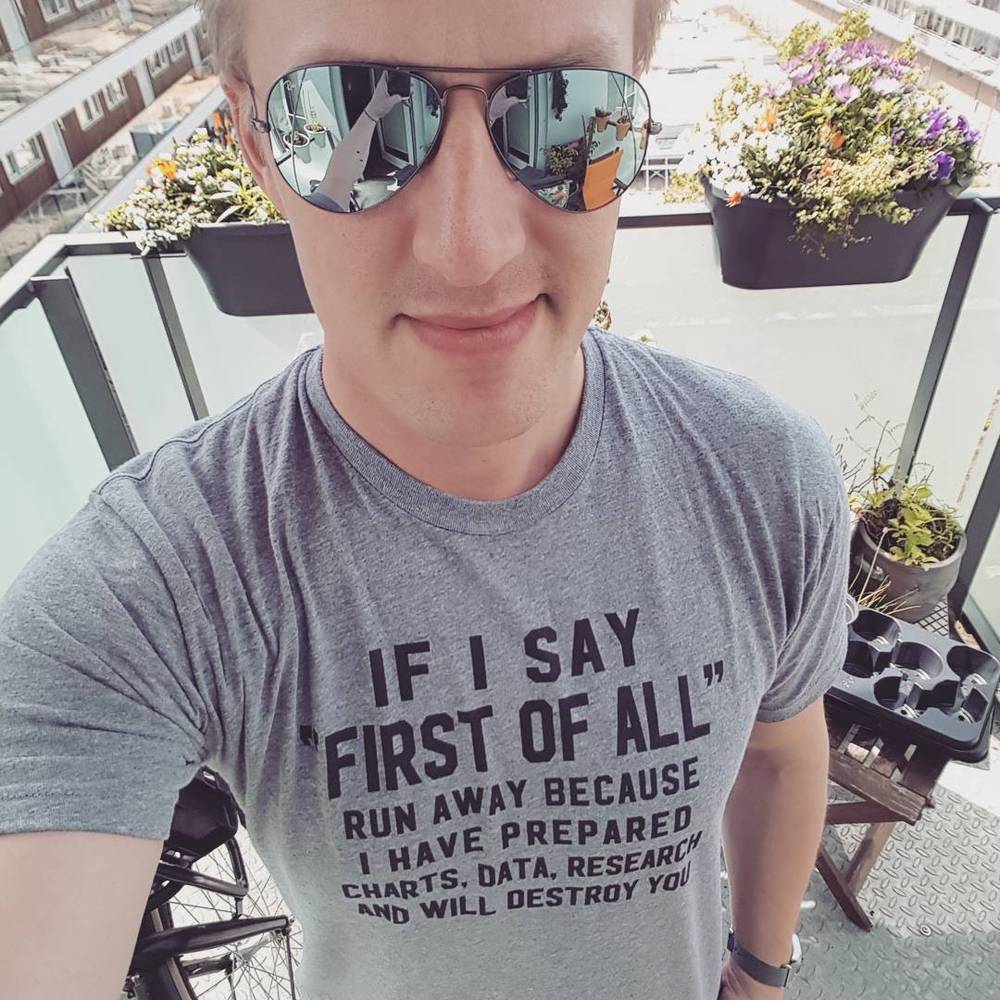 Close-up selfie of a man in mirrored sunglasses wearing a t-shirt reading "If I say 'first of all,' run away because I have prepared charts, data, research and will destroy you."