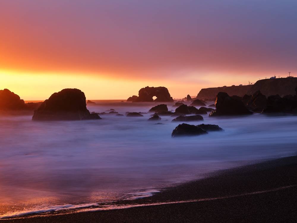 Arched Rock Beach in Sonoma Coast State Park near Carmet, California, at sunset. The naturally arched sea stack stands among scattered dark rocks in the surf, with long-exposure waves blurred into a soft mist across the beach. The sky glows in bands of orange, pink, and purple above the horizon, with a coastal headland and a few vehicles visible along the cliff to the right.