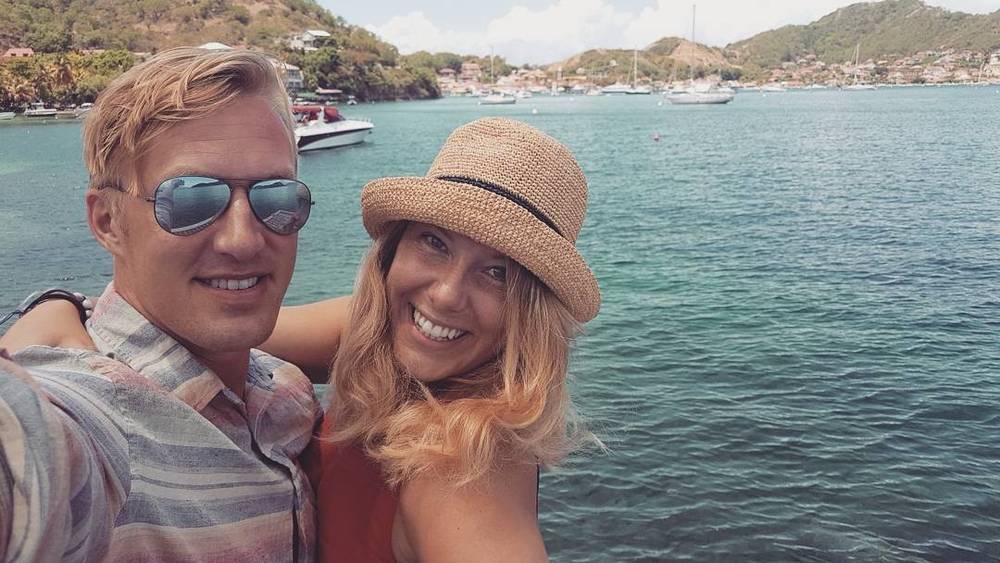 A couple taking a selfie on a boat with a bay and hills behind them.