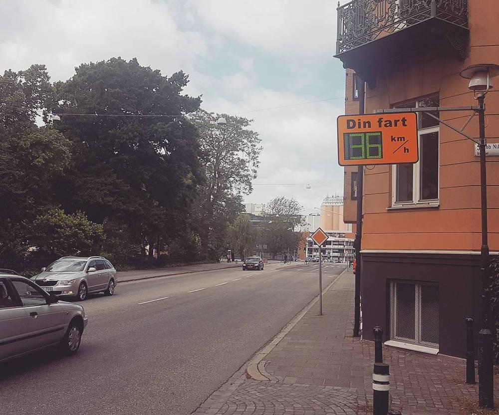 A quiet Swedish street with parked cars and trees, next to a digital speed sign showing a driver's current speed.