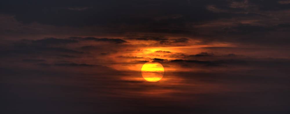The sun glows orange through layered clouds at sunset over a city skyline.