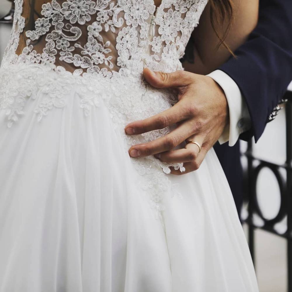 Close-up of a bride's lace wedding dress with a hand resting on it beside a groom's suited arm.