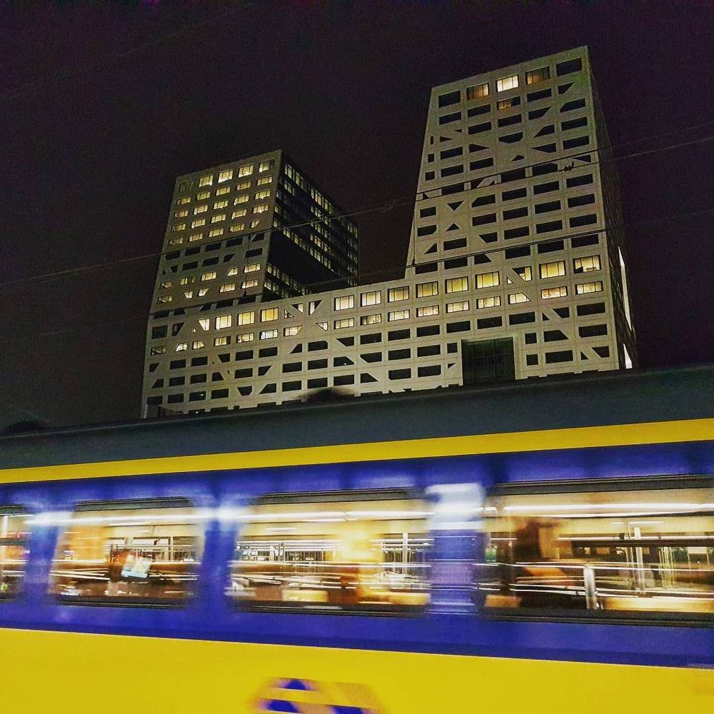 Two angular modern apartment buildings lit up at night beside a passing tram.