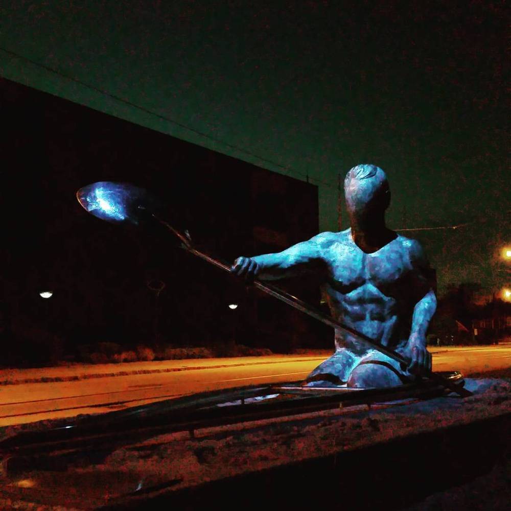 A blue-lit statue of a rower with an oar, illuminated at night beside a road.