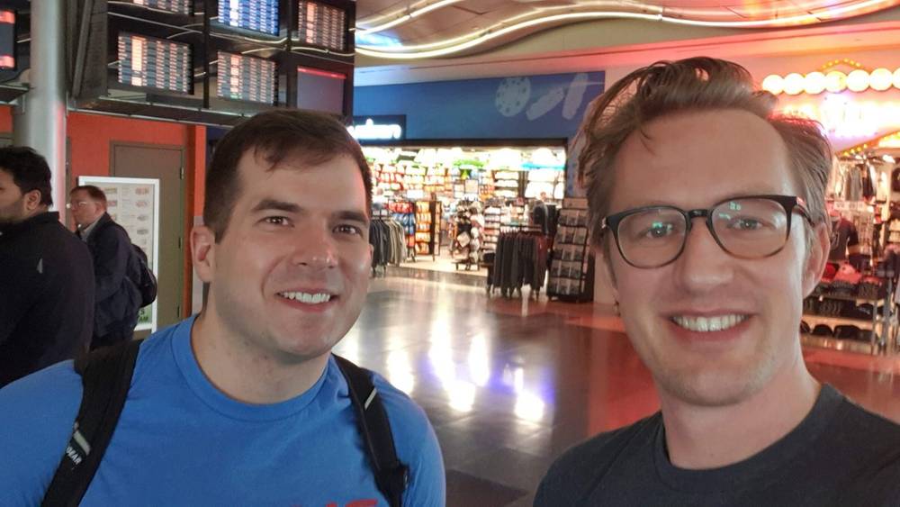 Two men smiling for a selfie in a venue with arcade games in the background.
