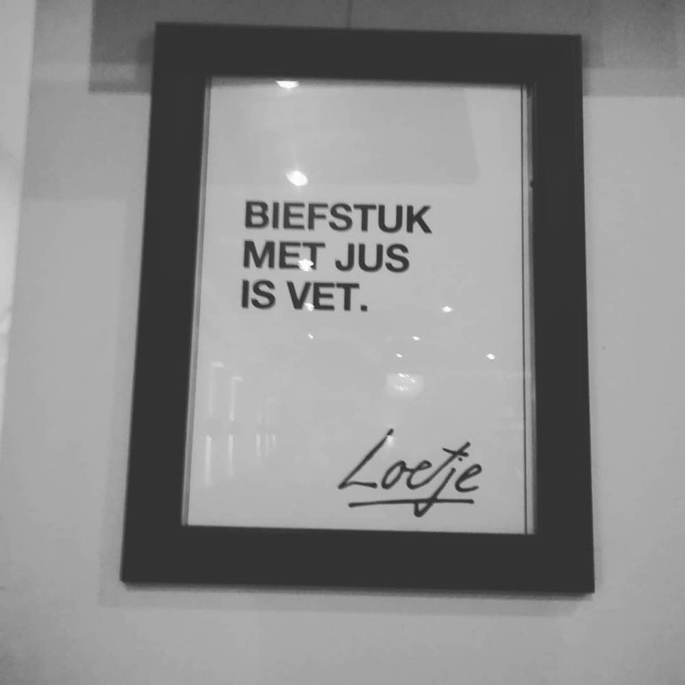 A framed black and white sign reading 'Biefstuk met jus is vet', signed Loetje.