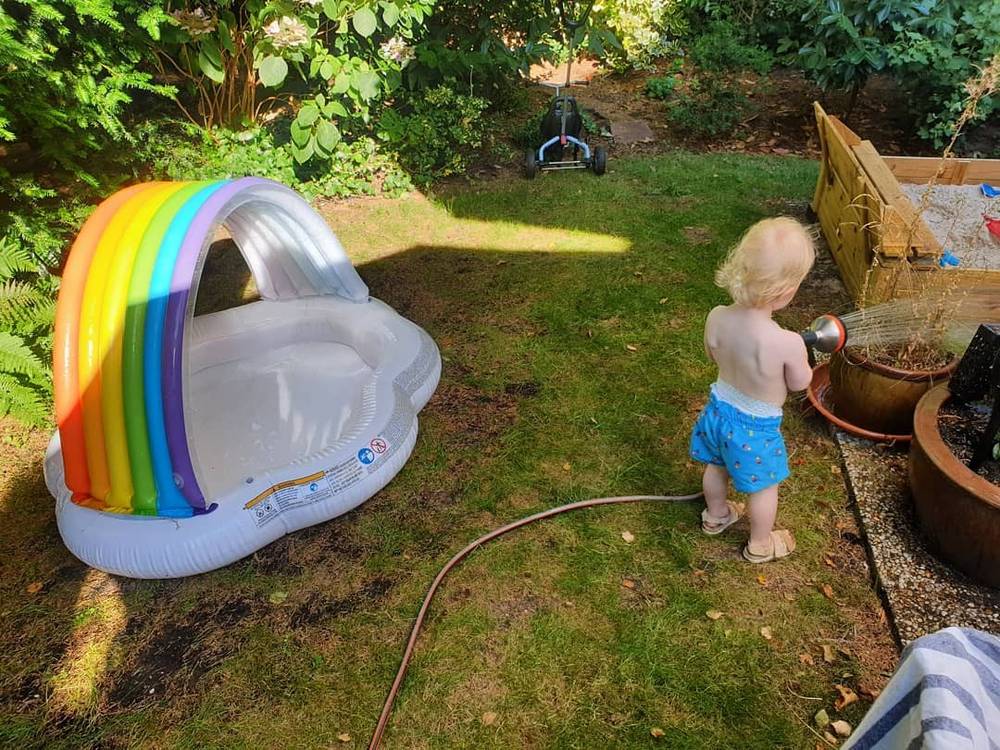 A toddler standing at a rainbow-striped inflatable pool canopy in a garden, with a hose running nearby.