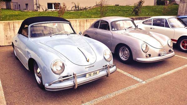 Two classic Porsche 356 cars, one light blue and one silver, parked side by side.