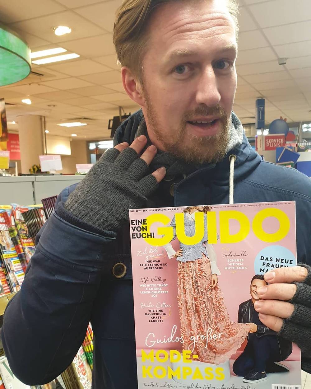 A man holds up a German magazine titled "Guido", smiling in a store.