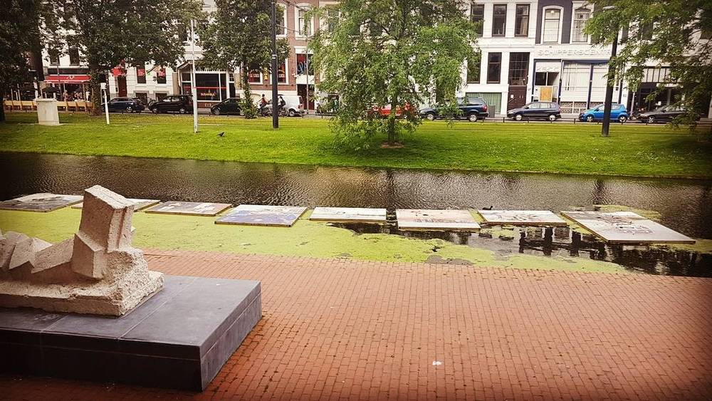 A park path along a canal with an abstract sculpture displayed on a plinth.