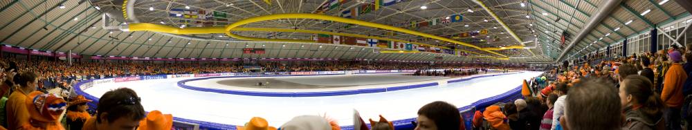 A packed indoor ice rink arena, spectators in orange cheering a speed skating event.