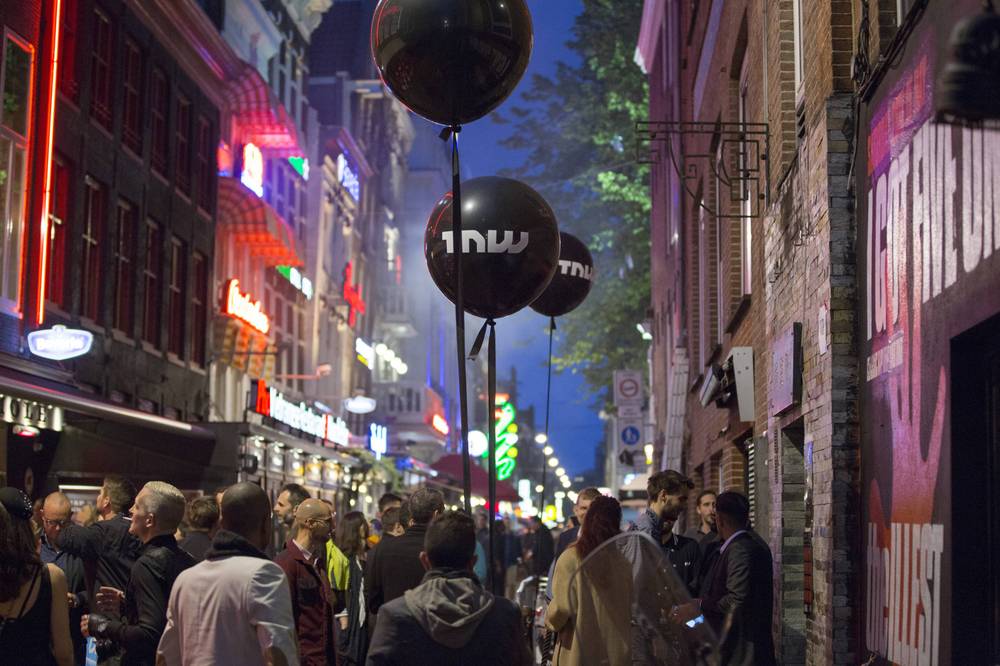 A crowd gathers on a neon-lit Amsterdam street beneath TNW balloons during an evening conference party.