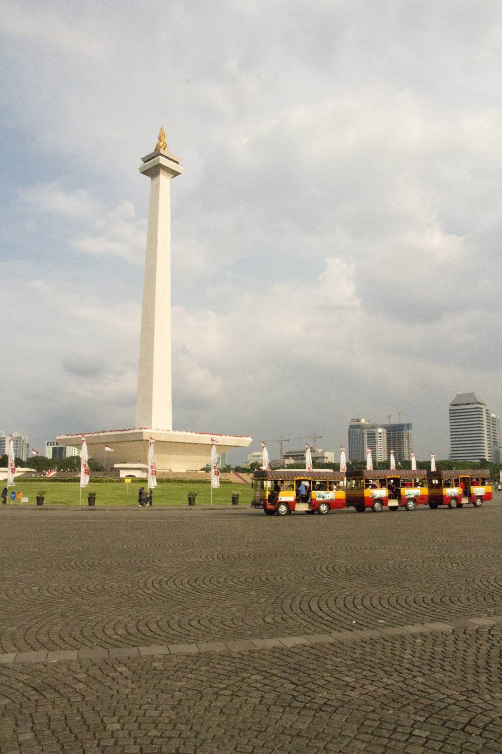 The National Monument tower stands above a paved plaza with small tourist trains parked nearby.