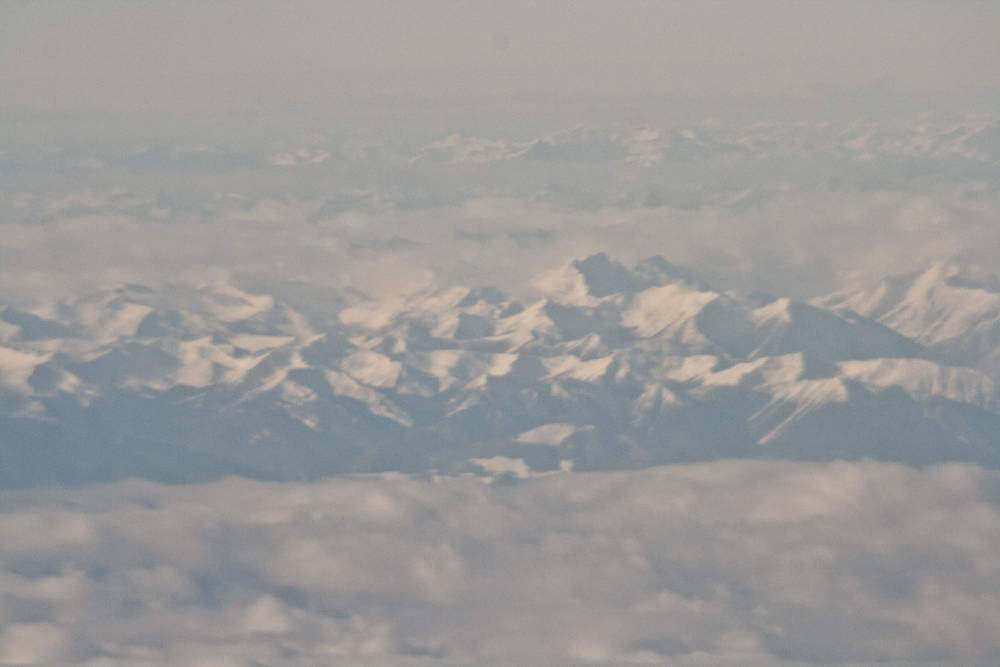 Snow-capped mountain peaks poke through a sea of clouds, seen from an airplane window.