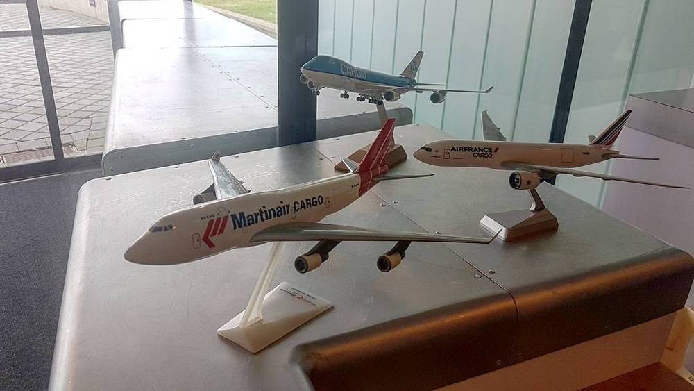 Model cargo airplanes displayed on a table, with a real jumbo jet visible through the window behind them.