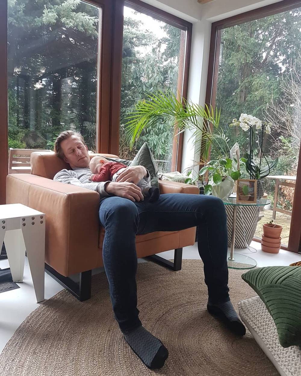 A man reclines on a couch with a baby beside large windows overlooking greenery.