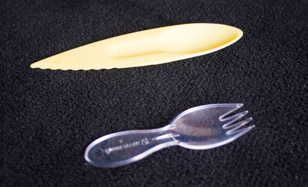 A novelty plastic utensil combining a spoon, knife and fork on a dark textured surface.