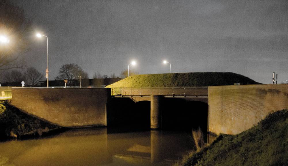 A concrete bridge crosses a dark canal beside a grassy embankment at night.