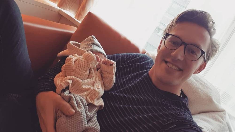 Selfie of a man smiling while holding a newborn baby wrapped in a blanket.