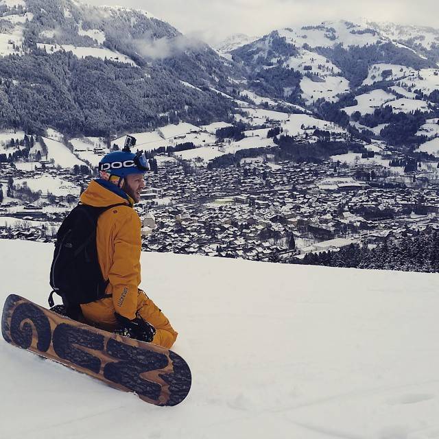 A snowboarder sits on a slope overlooking a snow-covered alpine town in the valley below.