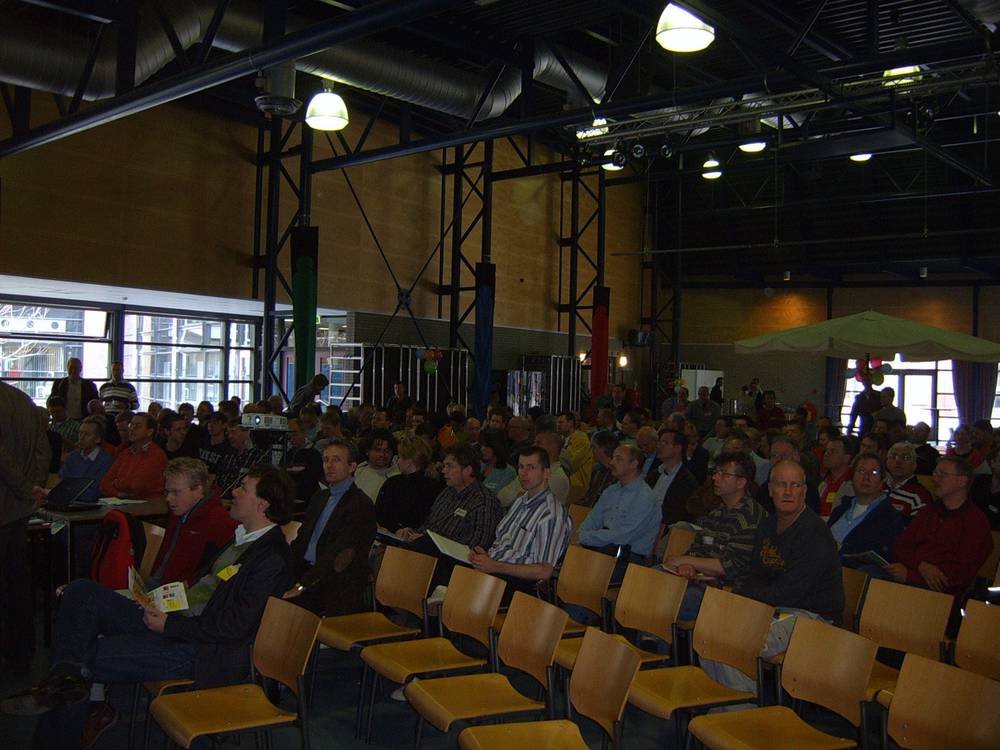 A large seated audience fills a conference hall with tall windows and steel roof trusses overhead.
