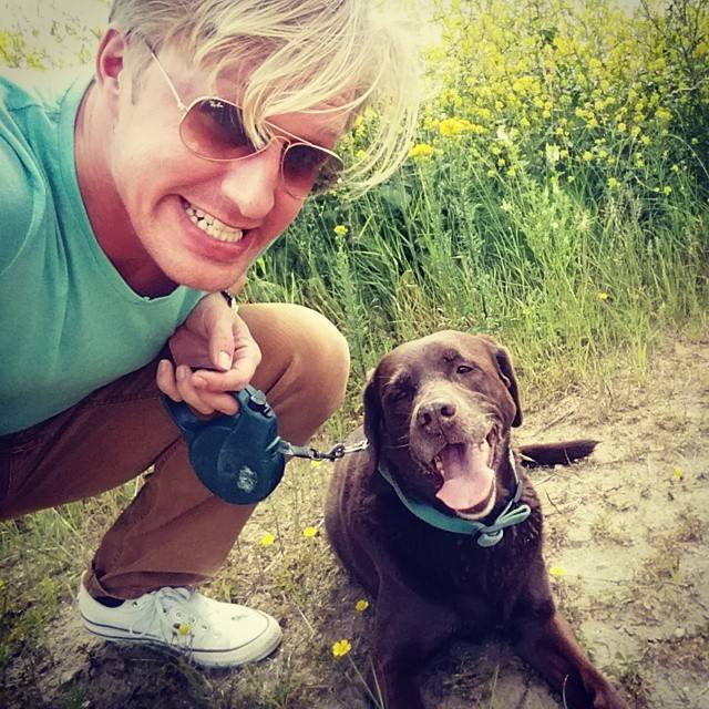 A man crouches beside a happy brown Labrador on a leash in a grassy field.